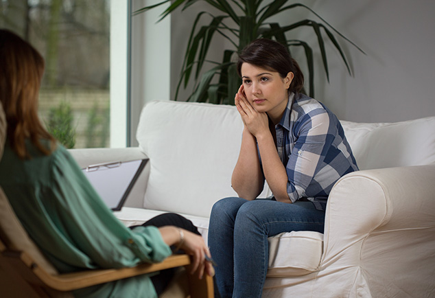 Woman sitting on sofa with her face resting on her hands looking at another woman sitting across her