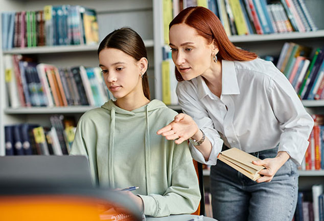 Student working on computer with teacher standing next to her helping